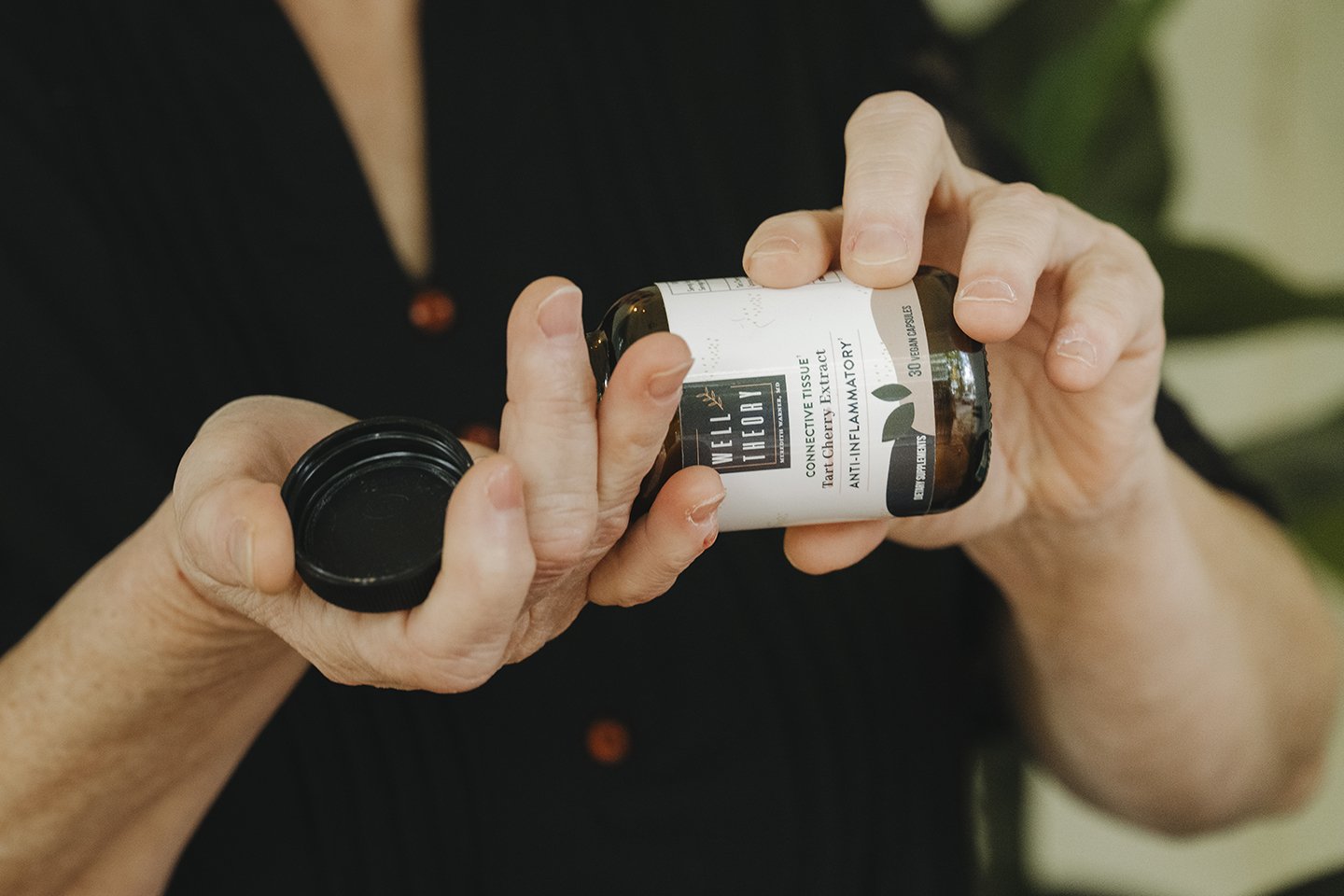A person pours capsules from a natural supplement bottle into their hand, with a plant blurred in the background.