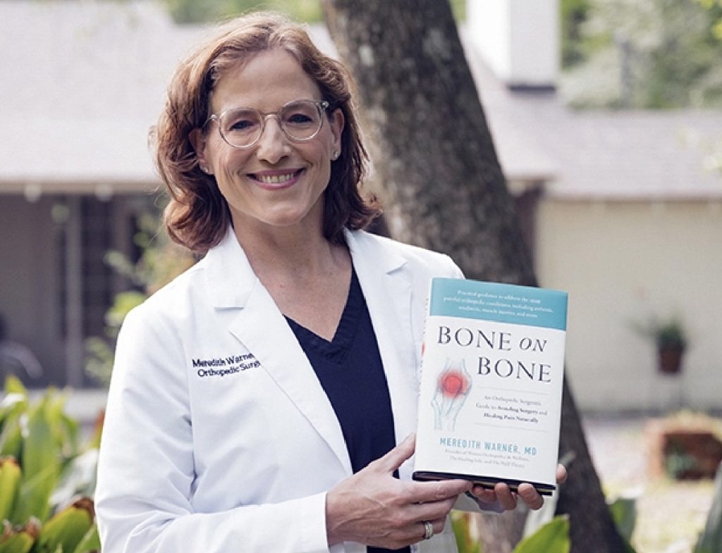 Smiling woman in a white lab coat outdoors, holding "Bone on Bone" supplements by Meredith Warner, MD.