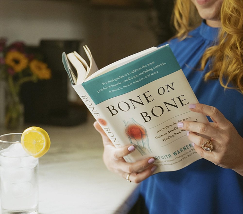 A person in a blue shirt reads "Bone on Bone" by Edith Warner beside iced water and surgeon-formulated supplements.