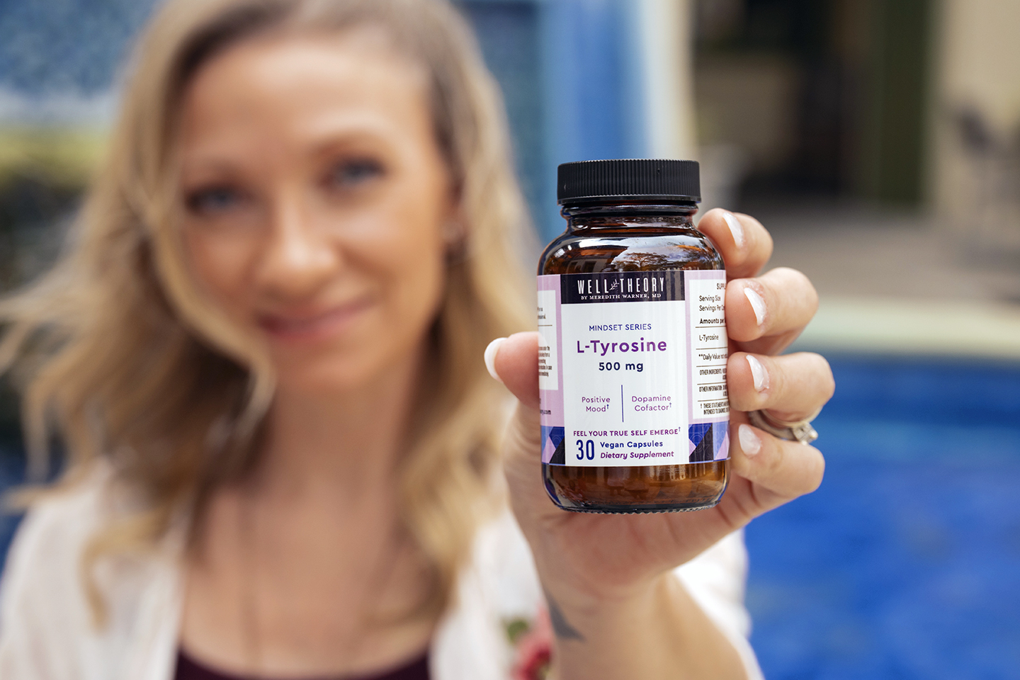 A person holds an “L-Tyrosine 500 mg” vegan supplement bottle with 30 capsules by a swimming pool.