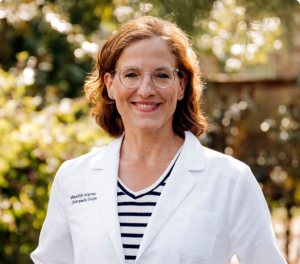 A smiling woman with brown hair and glasses in a white lab coat stands outdoors, promoting natural pain relief.