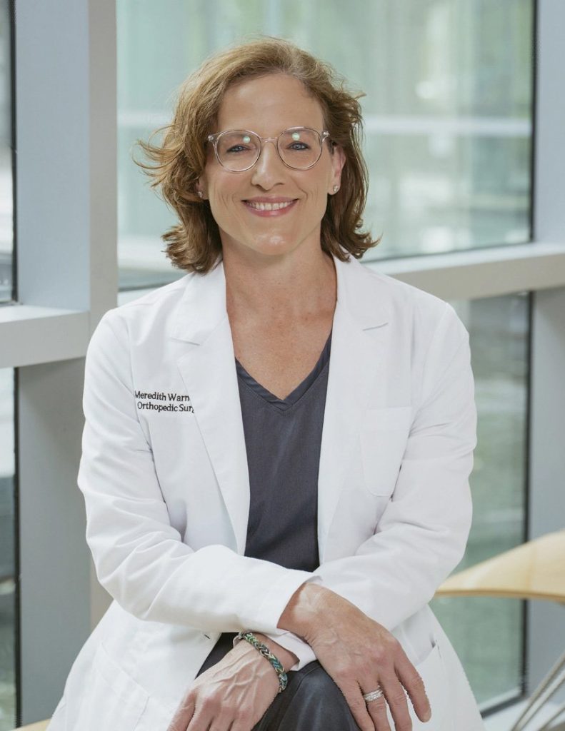 A woman in a white lab coat sits on a bench by large windows, smiling at the camera.