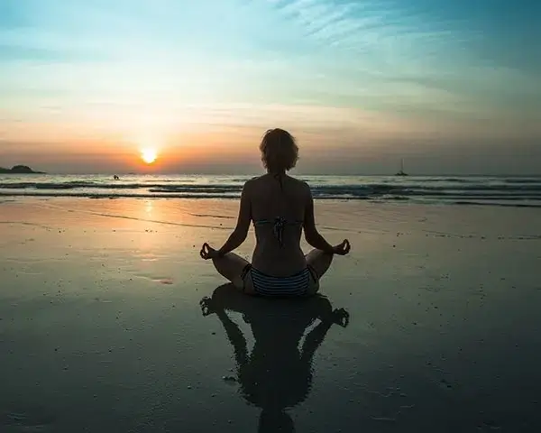 A person sits cross-legged on a beach at sunset, meditating and thinking about natural pain relief.